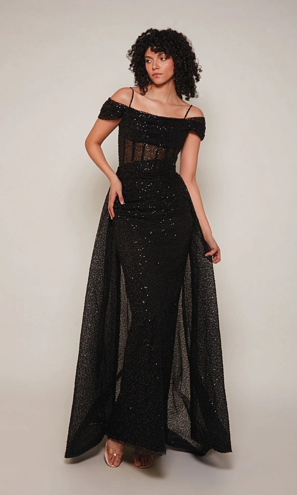 A woman poses against a plain light background wearing the Alyce 7101 long formal dress in black, featuring an off-shoulder design, spaghetti straps, sequined fabric, and sheer details.